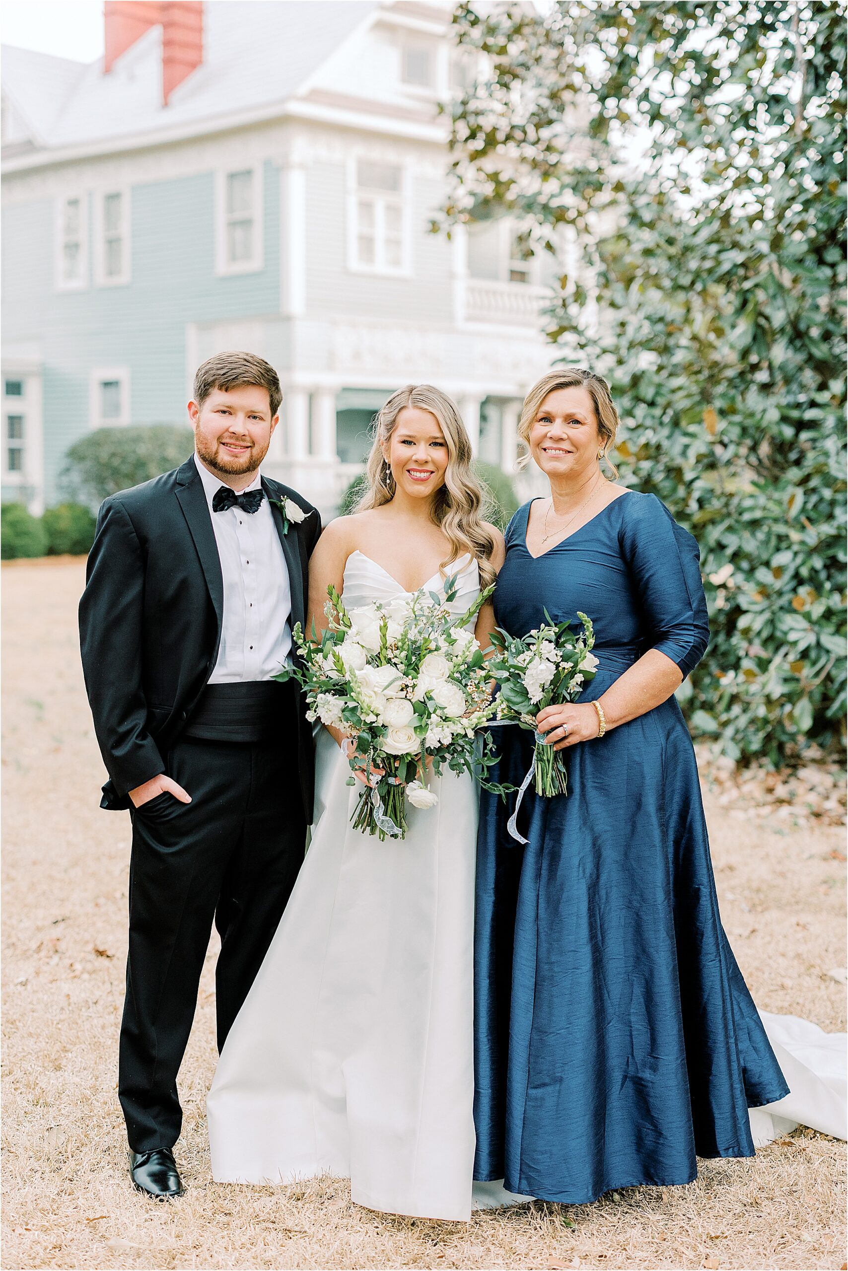 Mom, bride, and groom in front of a blue house surrounded by green trees.