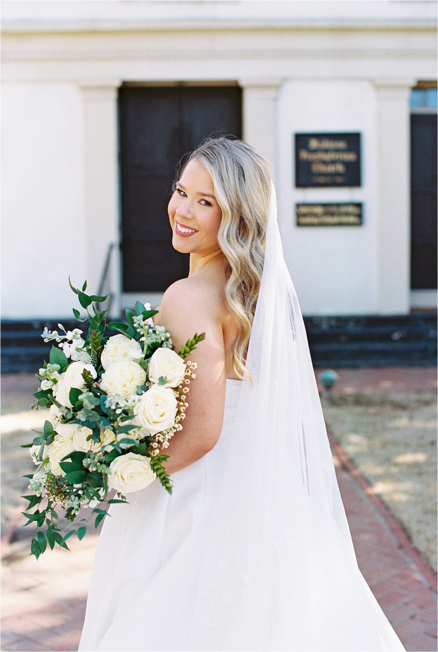 Bride in a white dress holding flowers standing in front of a white church.