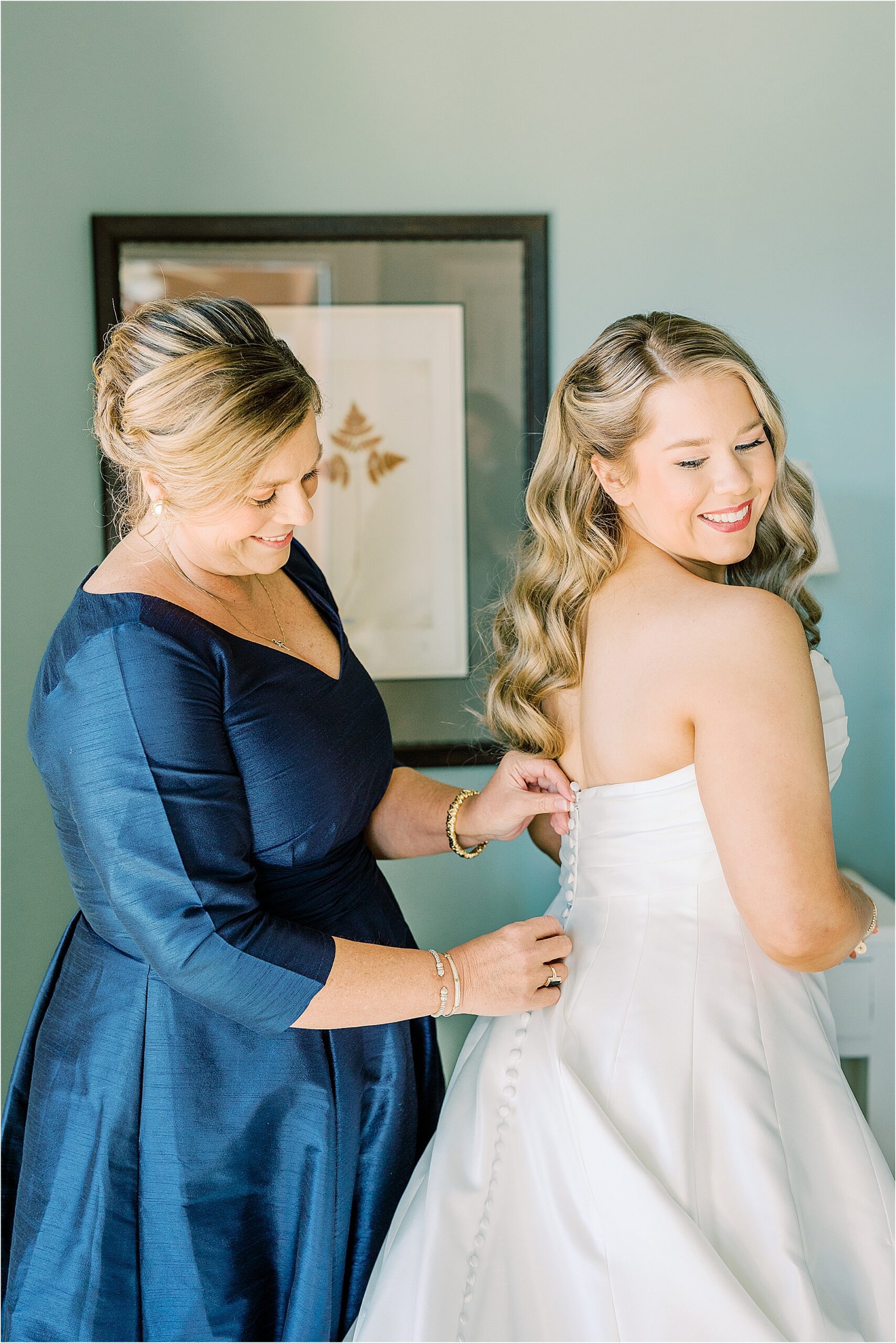 Woman in a blue dress buttoning the back of the bride's white wedding dress in a green room. 