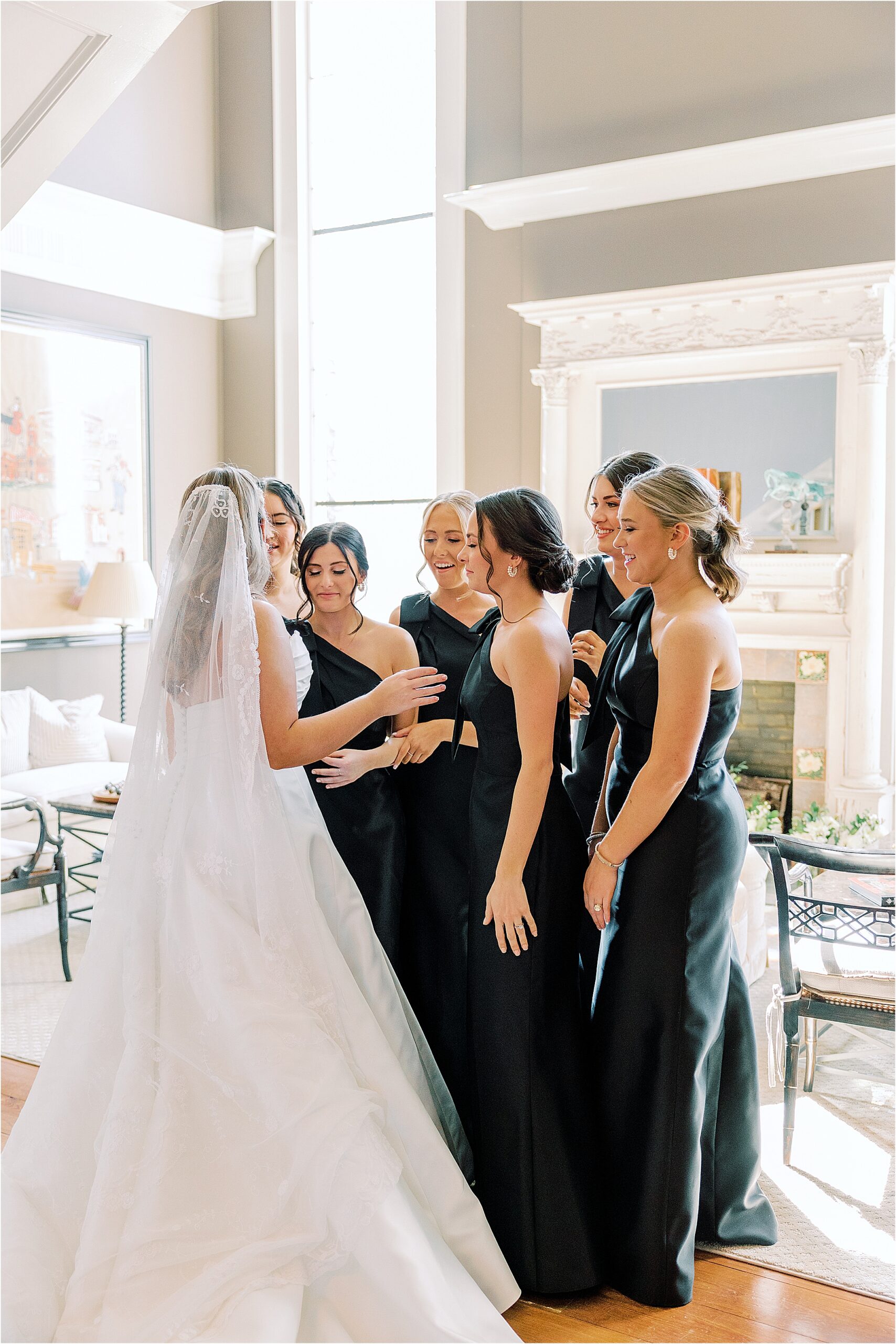 Bridesmaids in black dresses see the bride for the first time, standing in a tan room with big windows. 