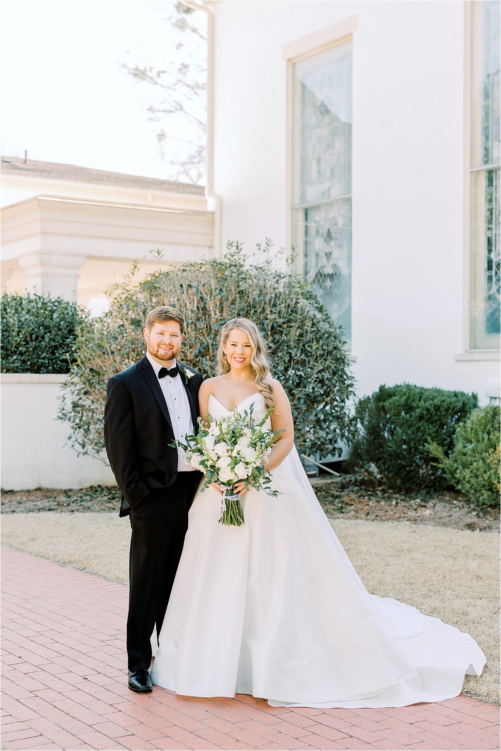 Bride in a white dress while holding flowers and the groom in a black tuxedo standing in front of a white church. 