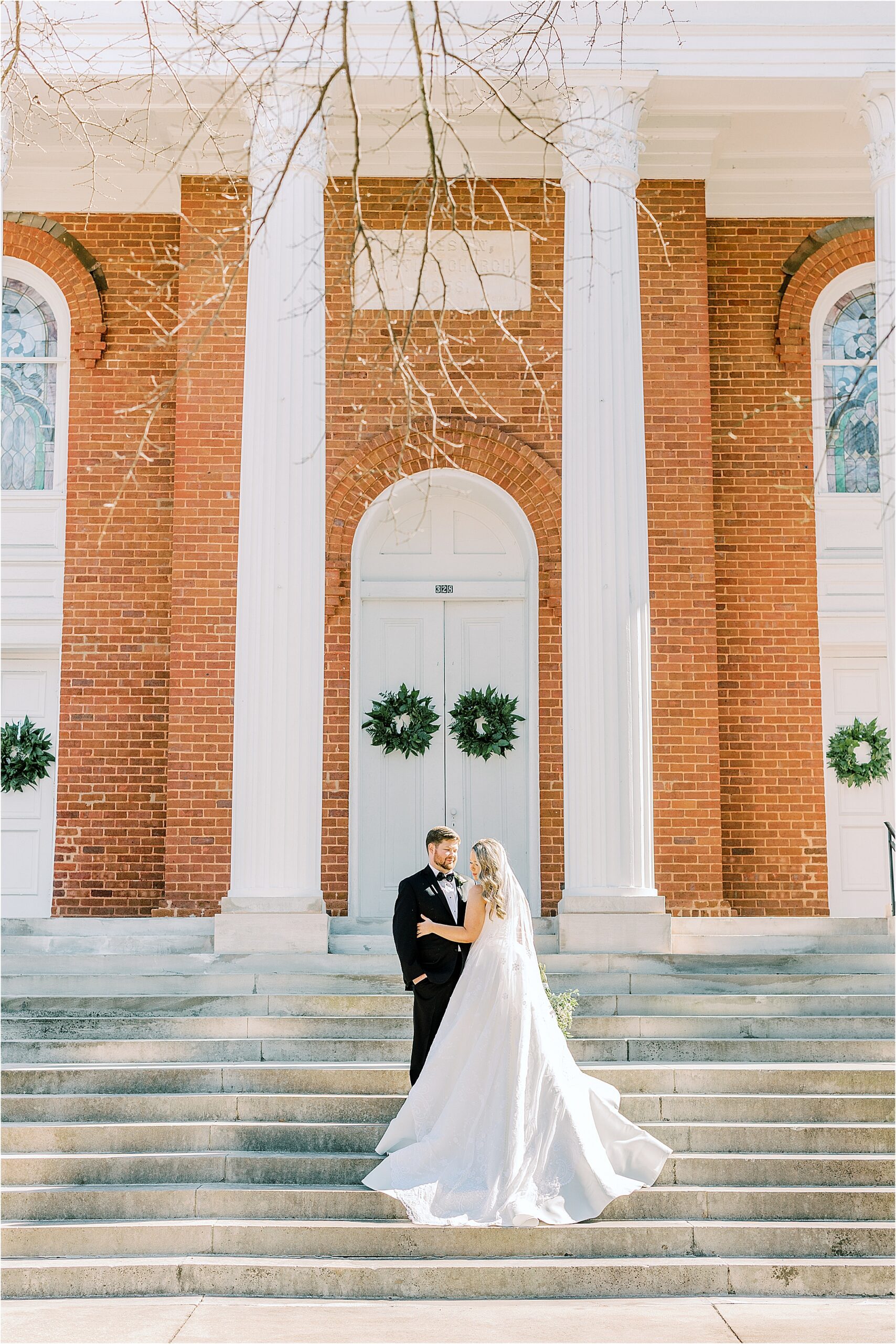 Bride in a white dress while holding flowers, and the groom in a black tuxedo is standing on the porch steps of a brick building with white doors. 
