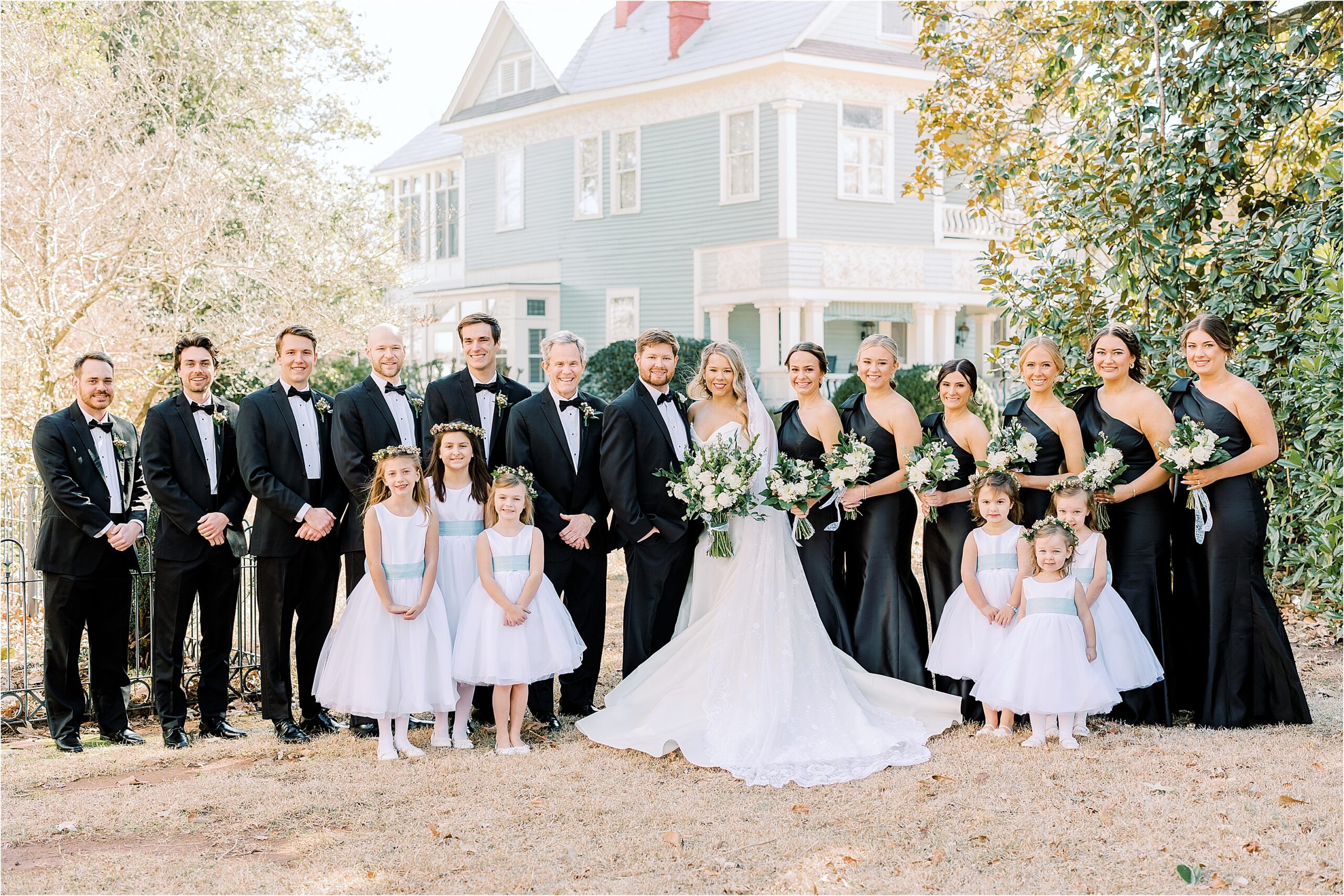 Bridal party in all black standing in front of a blue house sounded by trees. 